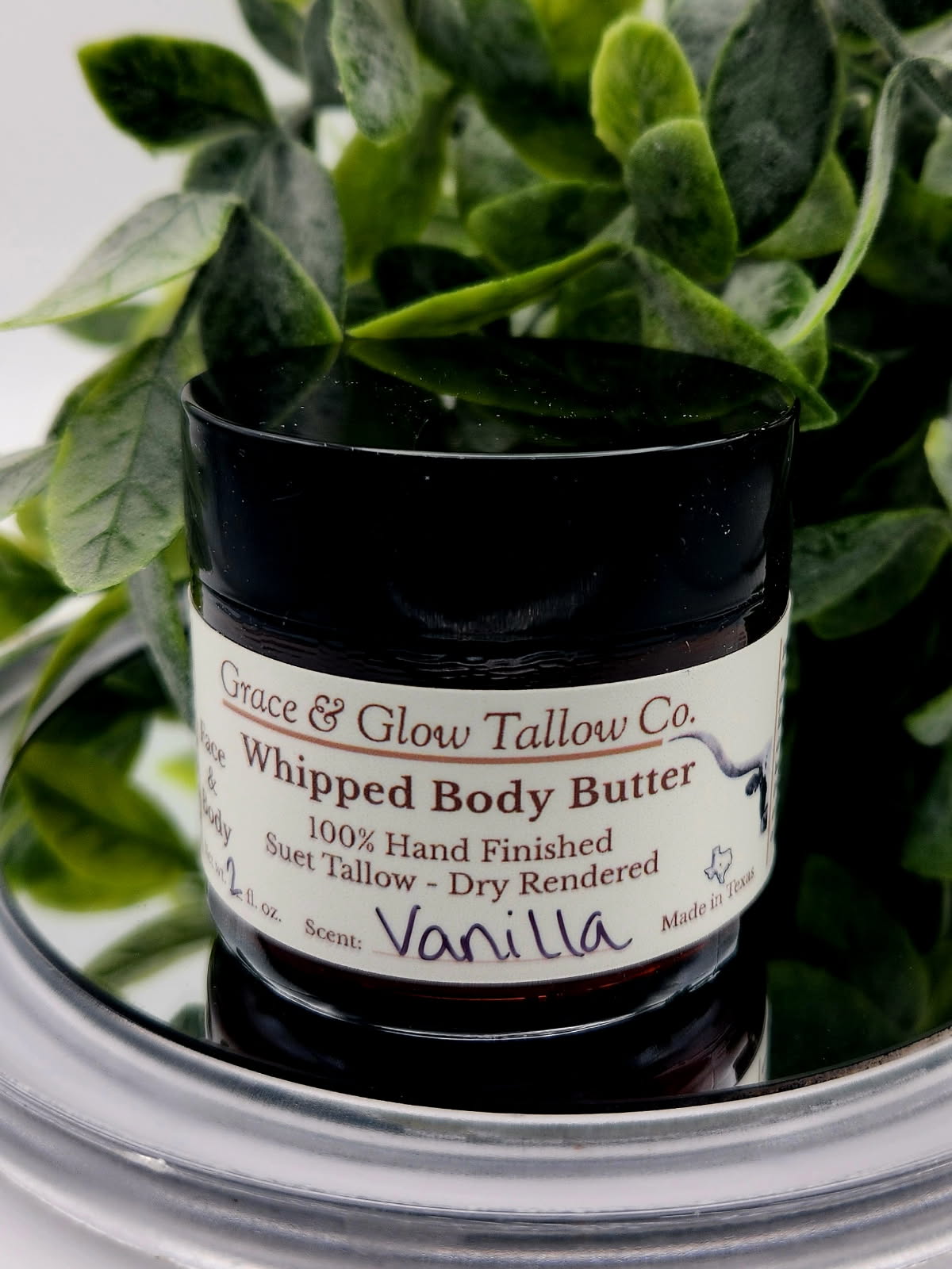 Jar of Grace & Glow Tallow Co. Whipped Body Butter with a vanilla scent, surrounded by green leaves.